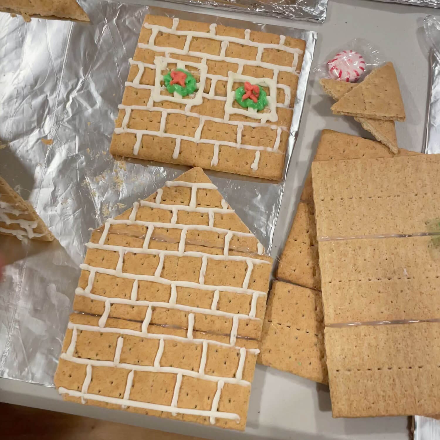 Graham cracker gingerbread house wall pieces piped with white royal icing in a brick pattern, with two small green and red wreath windows, ready to assemble.
