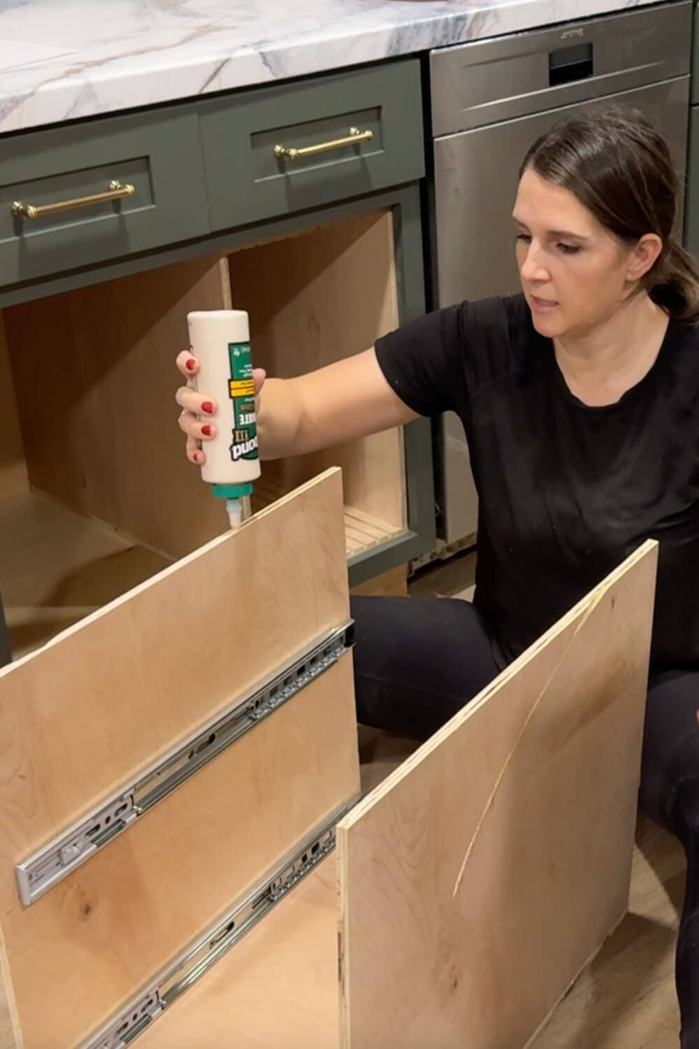 Woman applying wood glue along the edge of a plywood panel with installed drawer slides while building a custom pull-out organizer for glass food storage containers.