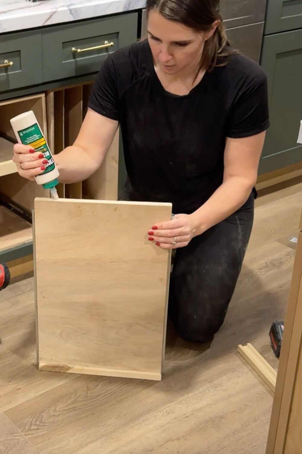 Woman applying wood glue along the edge of a plywood pull-out shelf before attaching a side rail for a DIY food storage container organizer.