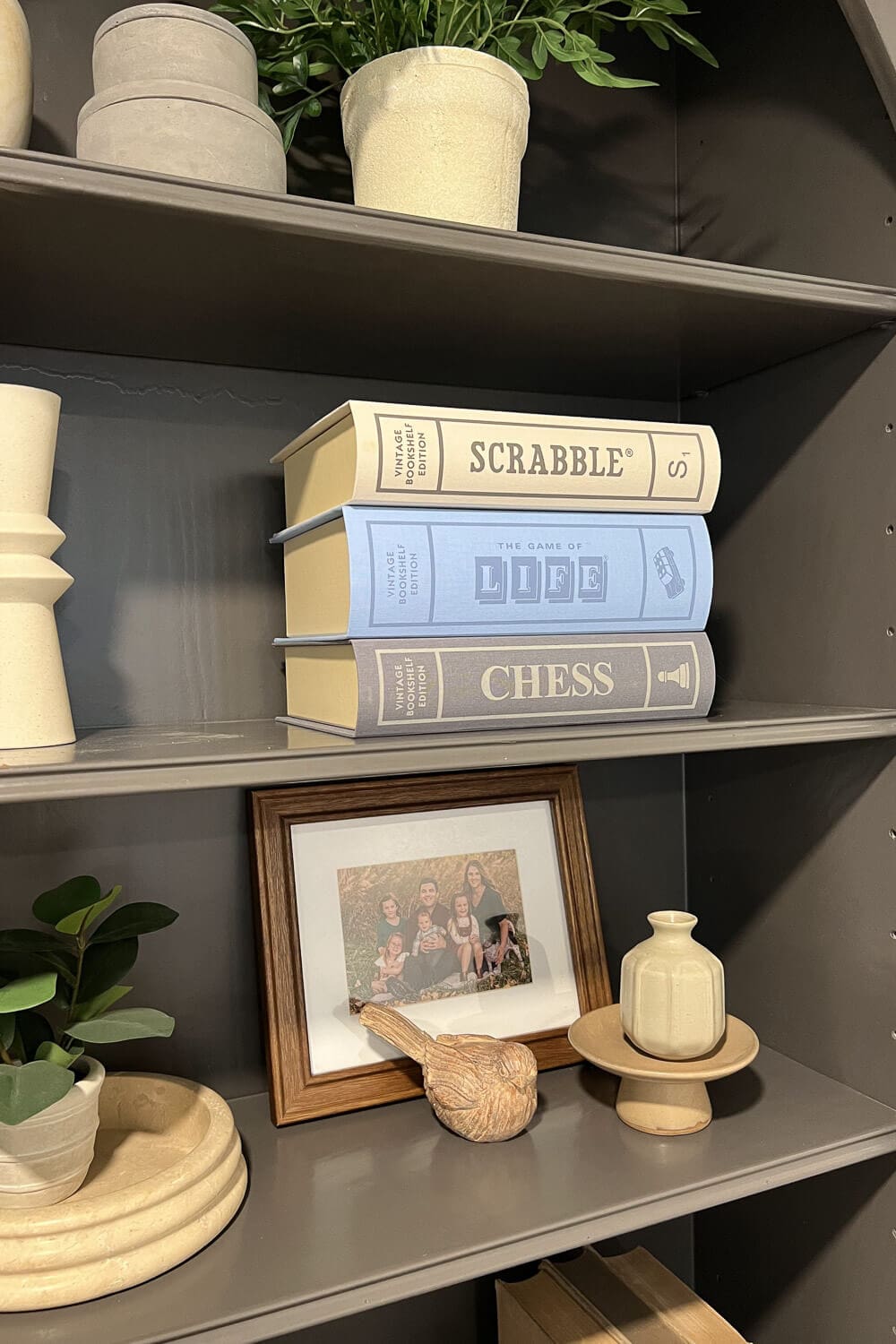Board Game Storage Ideas using vintage book-style board game boxes stacked on a shelf as decor, with Scrabble, The Game of Life, and Chess styled alongside plants and neutral home accents.