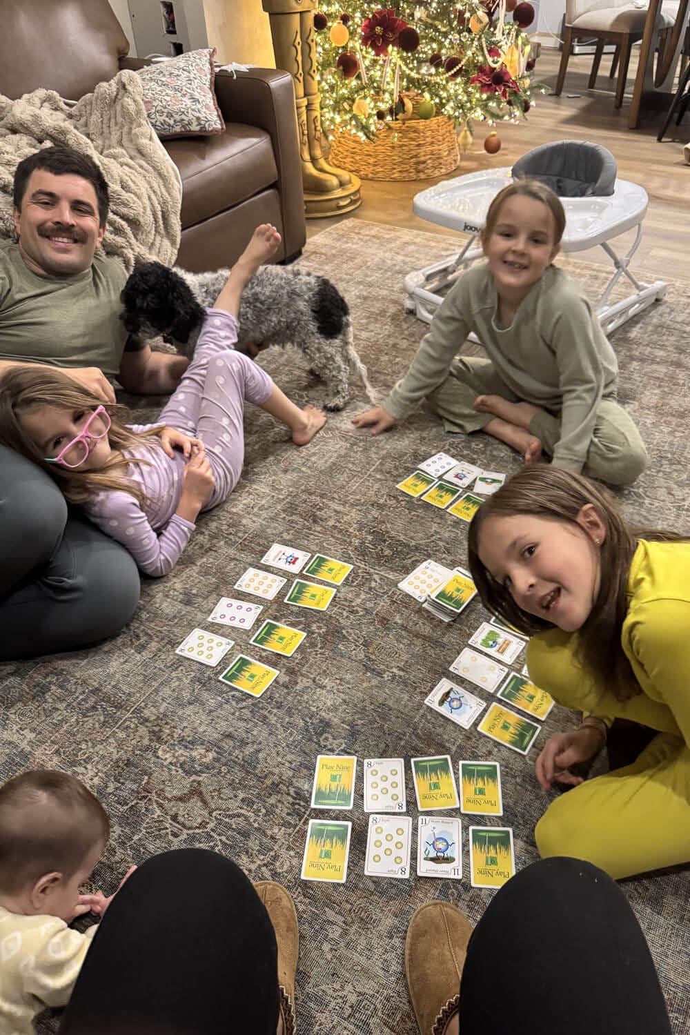 Family with small children playing a card game called Play 9 on the living room rug near the Christmas tree, showing.