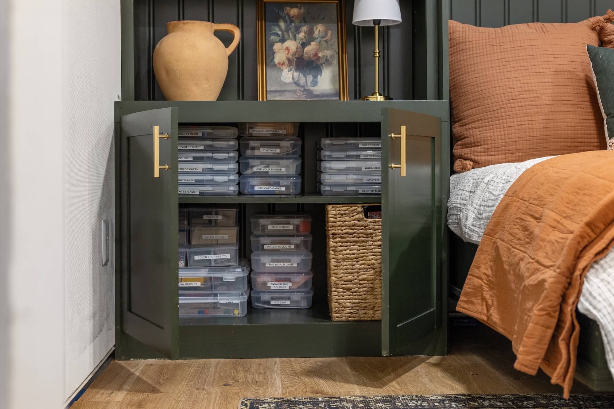 Board game storage inside a dark green built-in cabinet with open doors, showing neatly stacked and labeled clear plastic cases on two shelves with a woven basket beside them
