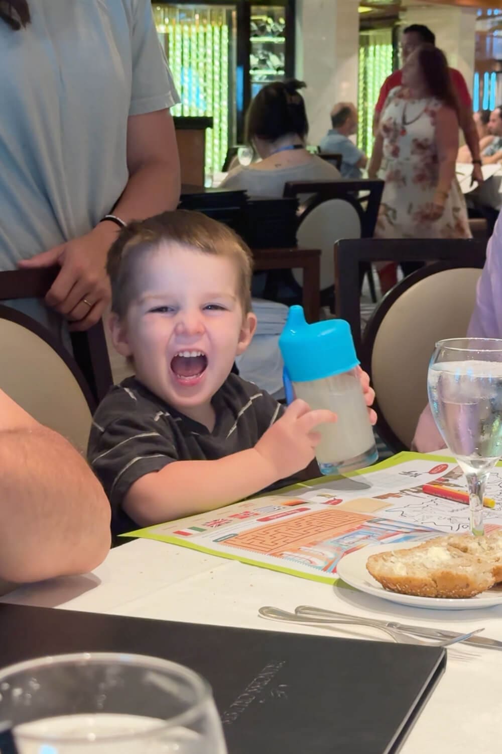 Toddler sitting at a cruise ship dining table holding a cup with a sippy cup lid topper, showing an easy cruise with kids tip for preventing spills during meals.