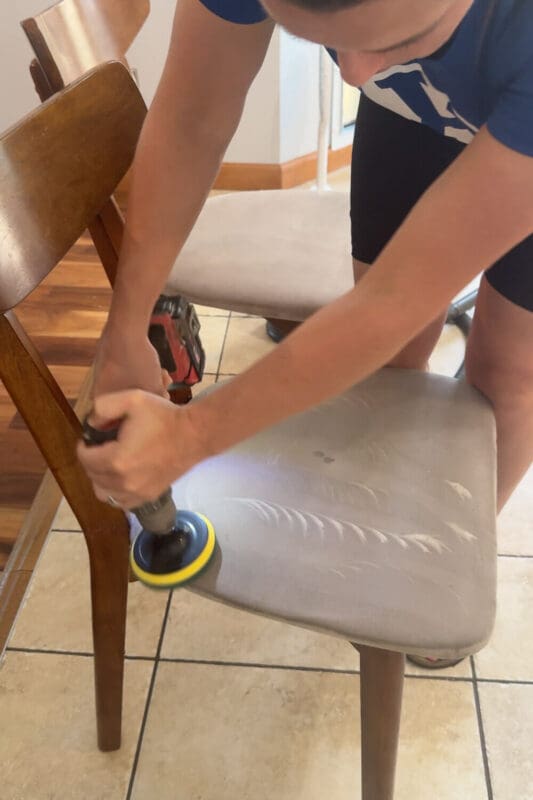 Using a drill brush to deep clean a stained upholstered dining chair seat, scrubbing out grime and set-in spots as part of a fabric chair cleaning tutorial.