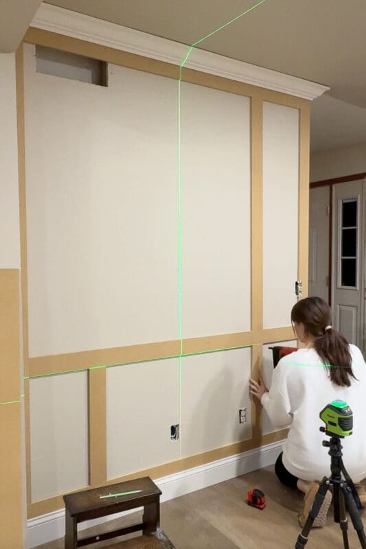 Building the DIY box molding wall trim grid by adding vertical trim pieces and smaller panels, using a laser level for even spacing.