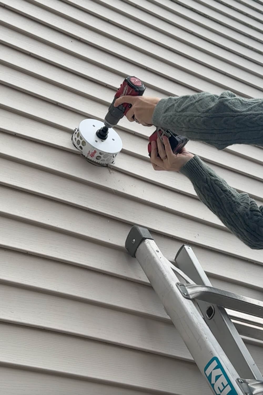 Close-up of hands using a drill with a hole saw to cut through exterior siding for the kitchen exhaust duct to vent outside.