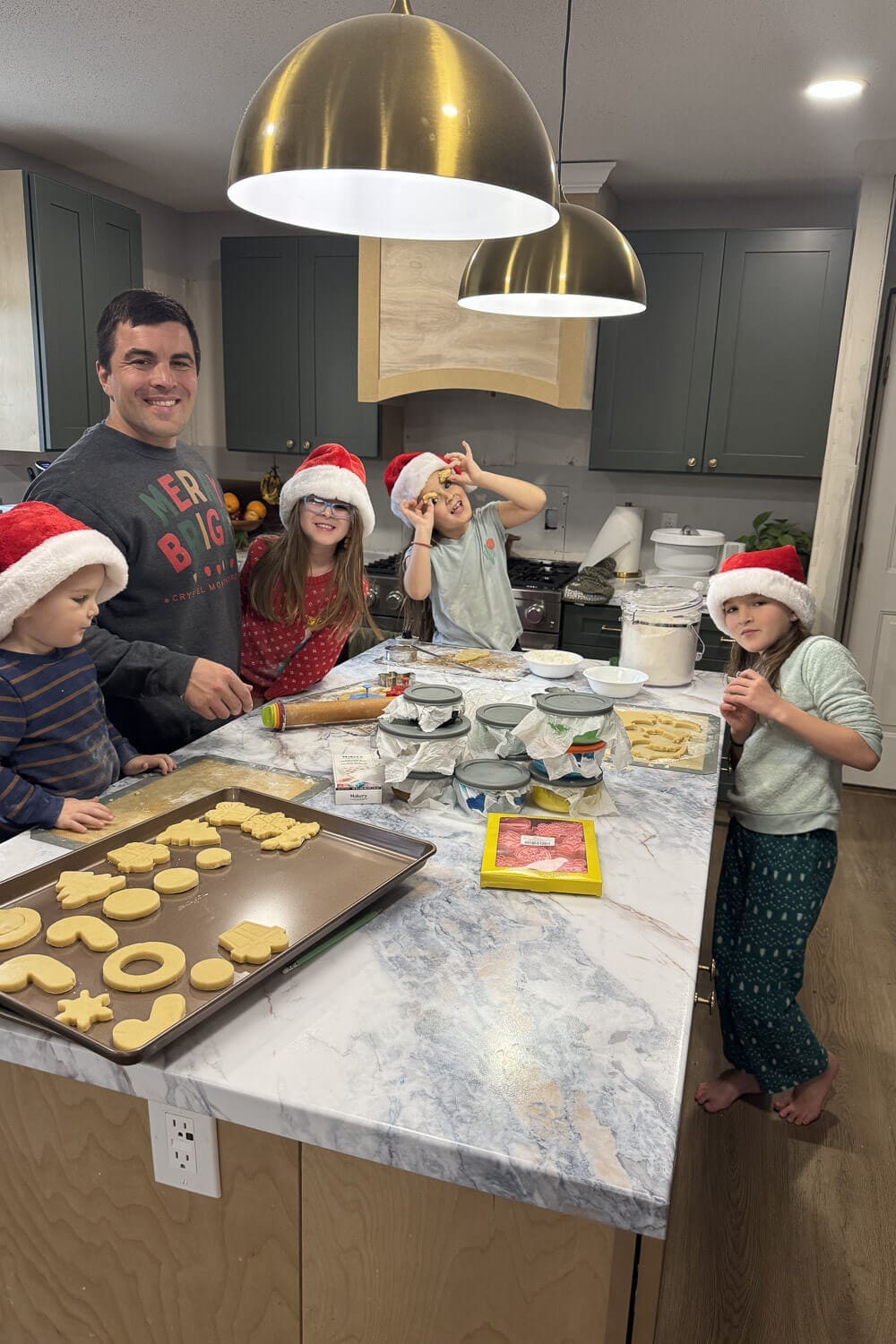 Family baking Christmas cookies around a DIY kitchen island with faux marble countertops, green cabinets, and brass pendant lights, with kids wearing Santa hats and cutout cookies on a baking sheet.