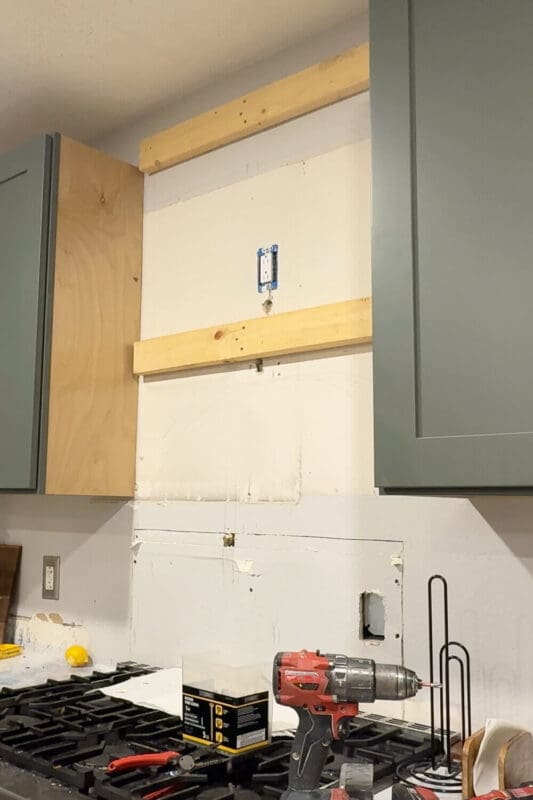 Kitchen wall above the stove with 2x4 blocking screwed into wall studs, creating sturdy support boards for mounting a DIY wood range hood cover frame between upper cabinets.