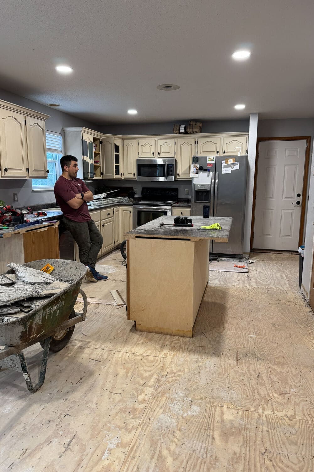 After cutting off the kitchen peninsula and removing the old flooring, the room is down to bare subfloor with the former peninsula cabinets moved to the center as a temporary island during demo.