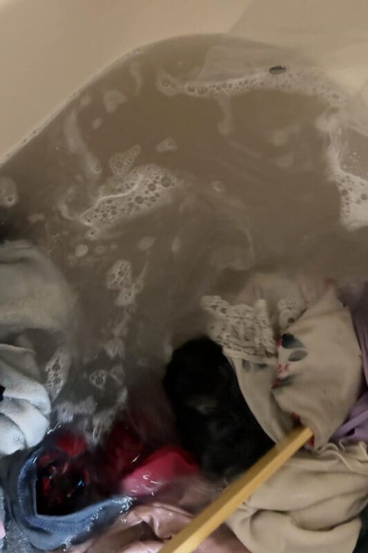 Stirring clothes in a bathtub during a deep cleaning as the water turns cloudy from gross detergent buildup and grime lifting out of fabrics.
