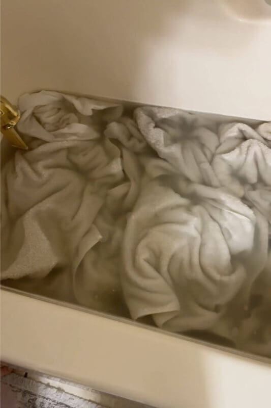 White towels soaking in a bathtub during laundry stripping, with cloudy water showing detergent buildup and residue being pulled out of the fabric.