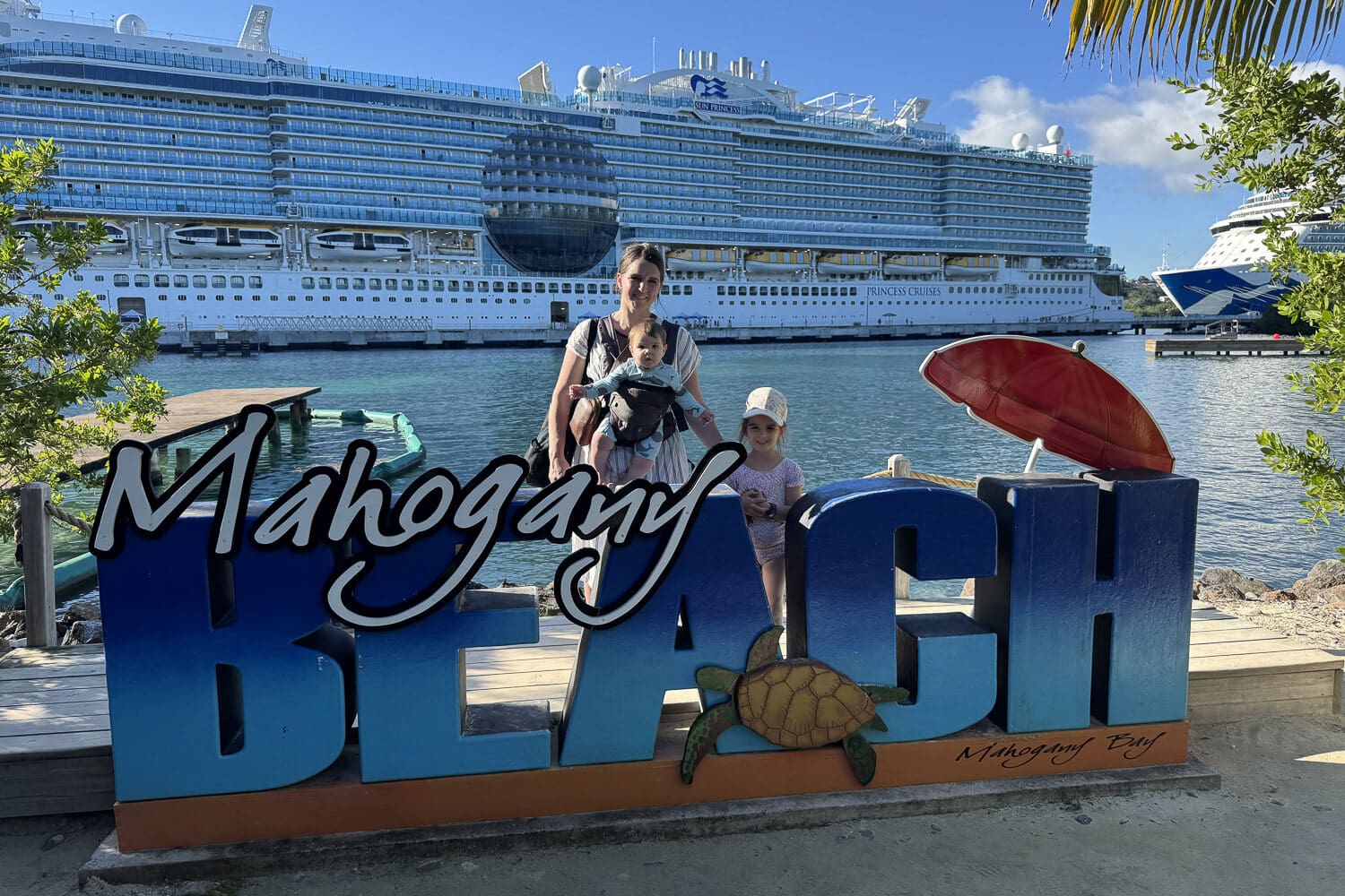 Mom wearing a baby in a carrier with a young child standing beside her at the Mahogany Beach sign in Mahogany Bay, Roatán, with a Princess Cruises ship docked in the background on a cruise with kids.