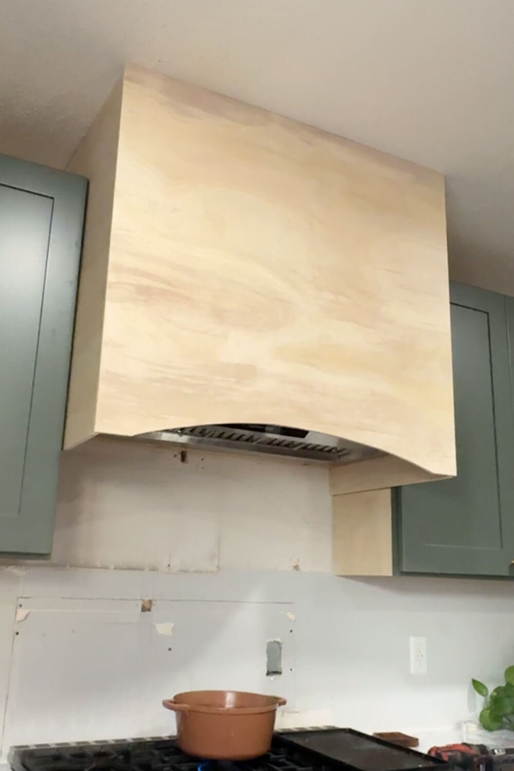 DIY range hood cover fully wrapped in plywood with a smooth arched front, installed between upper kitchen cabinets above the stove.