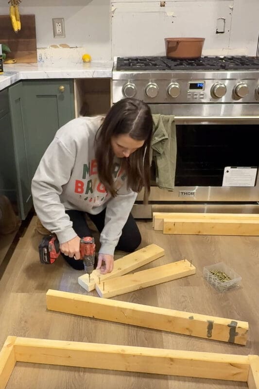 DIY builder kneeling on the kitchen floor using a cordless drill to predrill and screw corner blocks onto cut lumber pieces, prepping the wood frame for a DIY range hood cover above a stainless gas range.