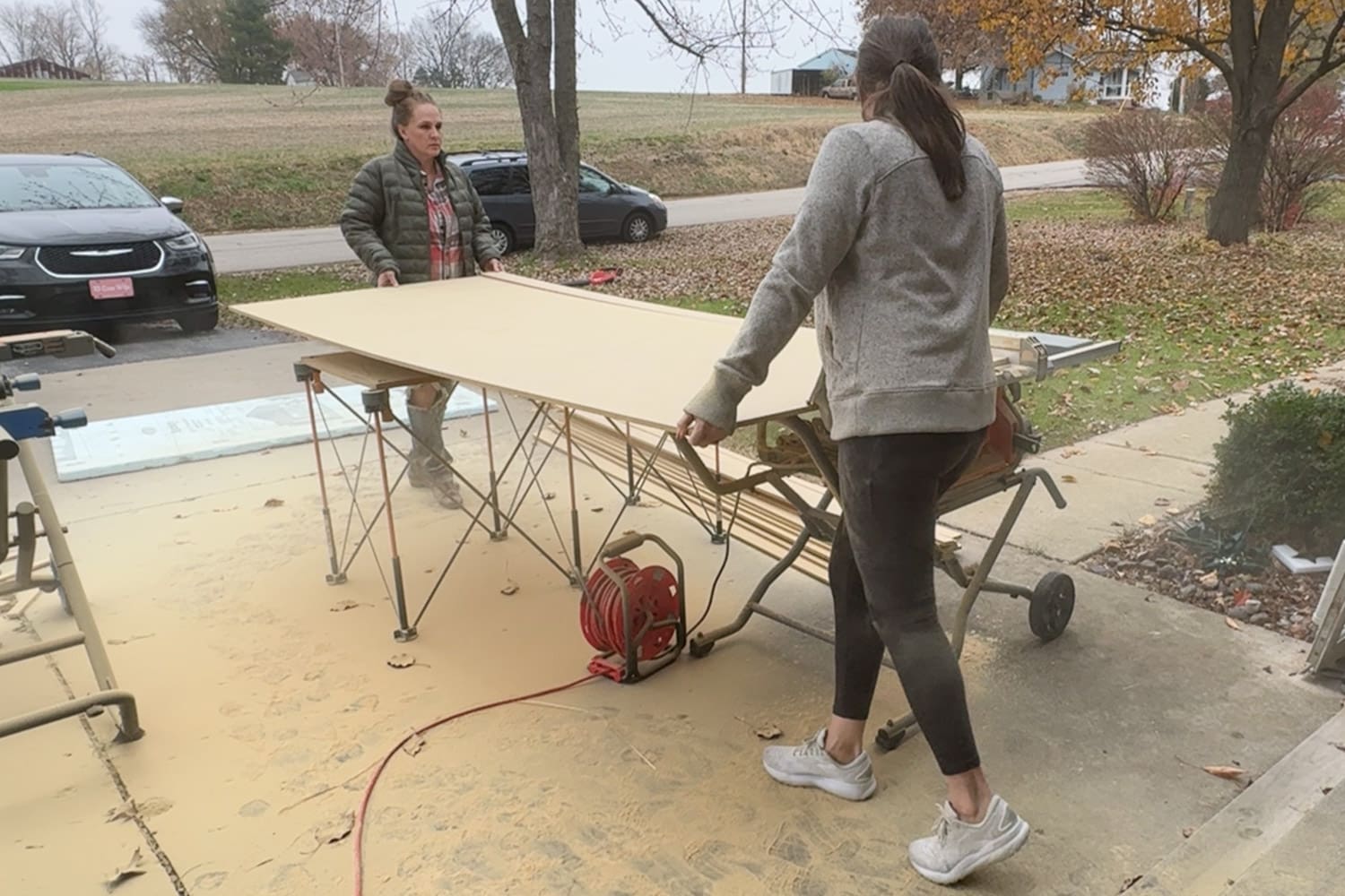 Two women carry a large 1/2-inch MDF sheet outside near a table saw and sawhorses to rip it into DIY trim boards wall trim.