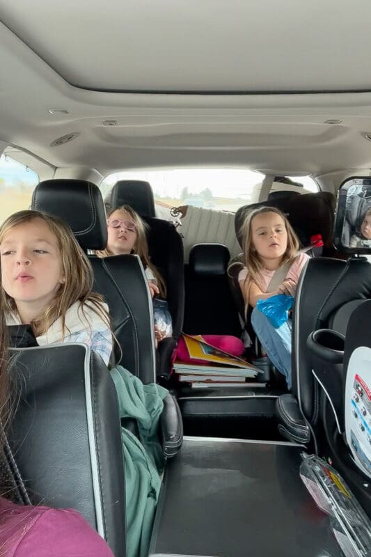 Kids strapped into car seats in the back of an SUV during a road trip with kids, with books and travel gear in the middle row, showing real-life road trip essentials for families.
