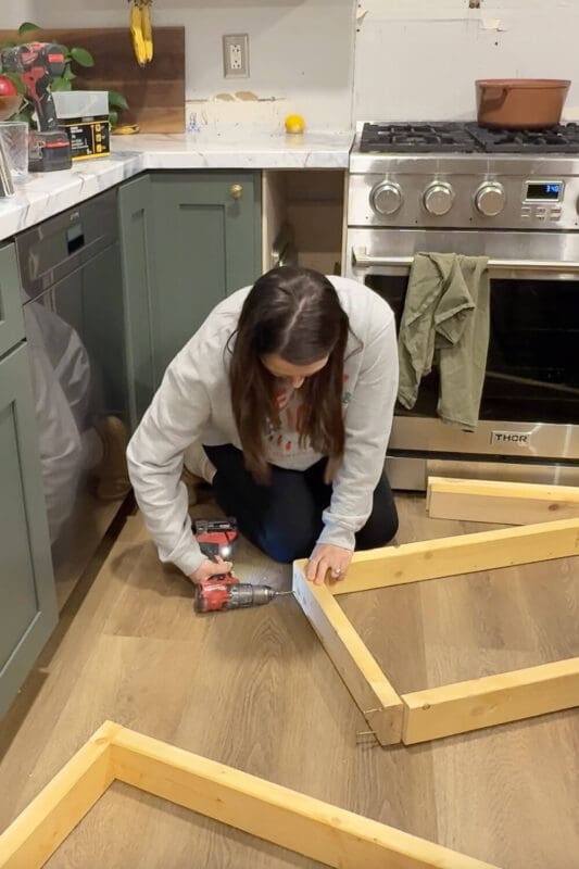 DIY builder assembling the wood range hood cover frame by drilling screws through a corner block to join two boards, forming a rectangular frame on the kitchen floor in front of green kitchen cabinets and a stainless steel stove.