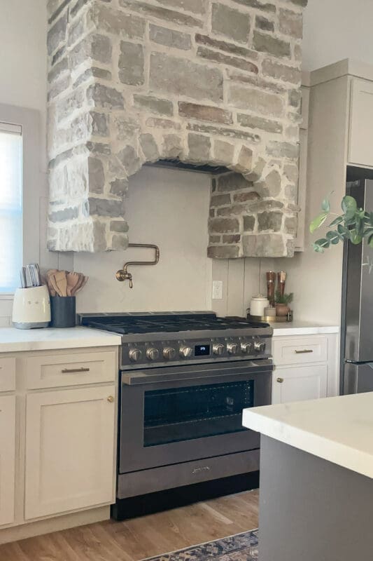 Stone-veneer range hood surround with an arched opening over a stainless range, paired with white cabinets, a brass pot filler, and simple neutral kitchen styling.