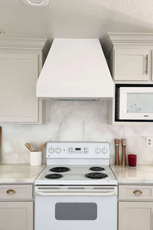 White painted wood range hood cover with a clean, tapered shape between white shaker cabinets, styled above a white stove with a light marble-look backsplash.