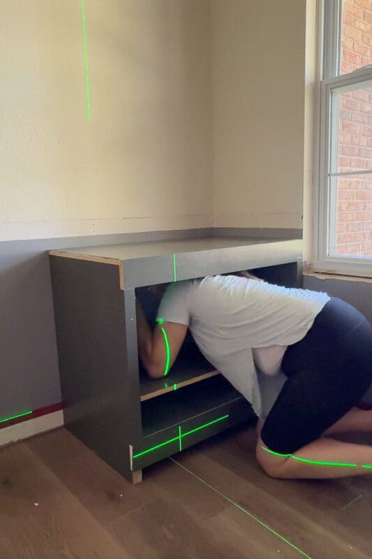 Attaching a finished DIY base cabinet to the wall using a laser level for alignment in a built-in cabinet project.