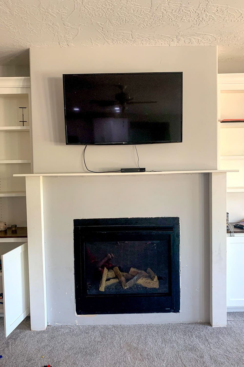 Dated builder-grade white fireplace with stepped drywall surround, thin mantel shelf, TV mounted with visible cords, white built-ins on sides