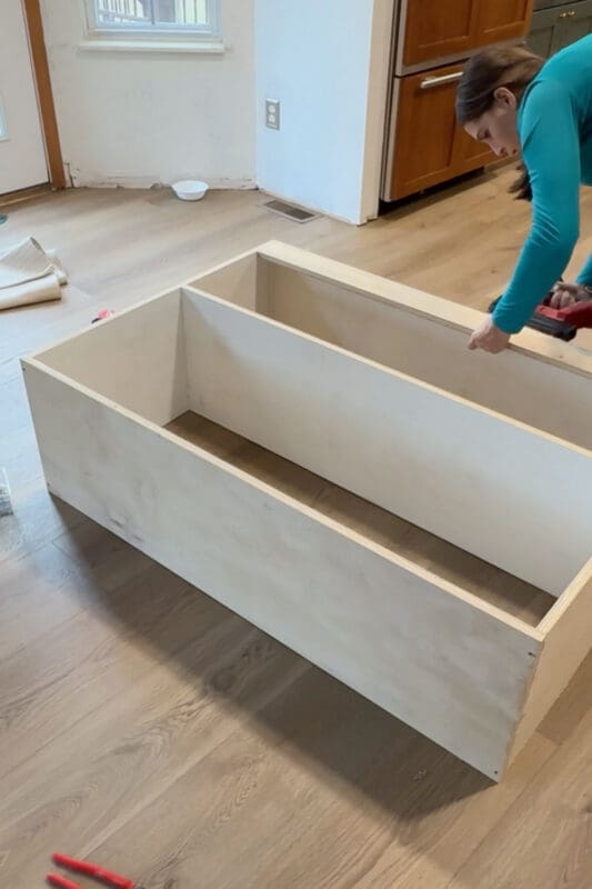 Crystel using a brad nailer to assemble a plywood box on the floor, building the floating shelf unit that will hang between the DIY built-in bookshelves in our home library.