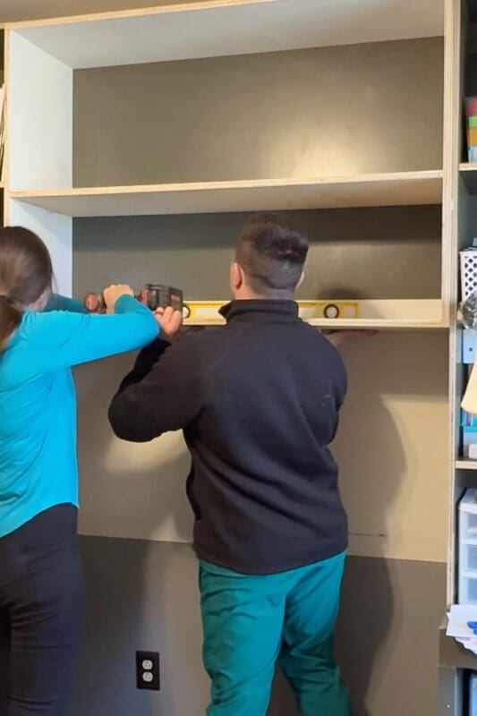 Crystel and a helper holding a level against the floating shelf while driving screws through the rear stretcher into wall studs, installing the last of our DIY built-in bookshelves.