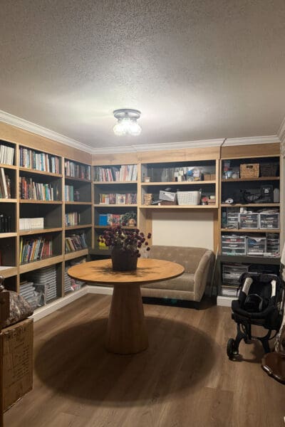Home library after moving in, with floor-to-ceiling built-in bookshelves filled with books and storage bins around a round table and small loveseat, showing new crown molding and baseboards with the shelf edges still raw wood.