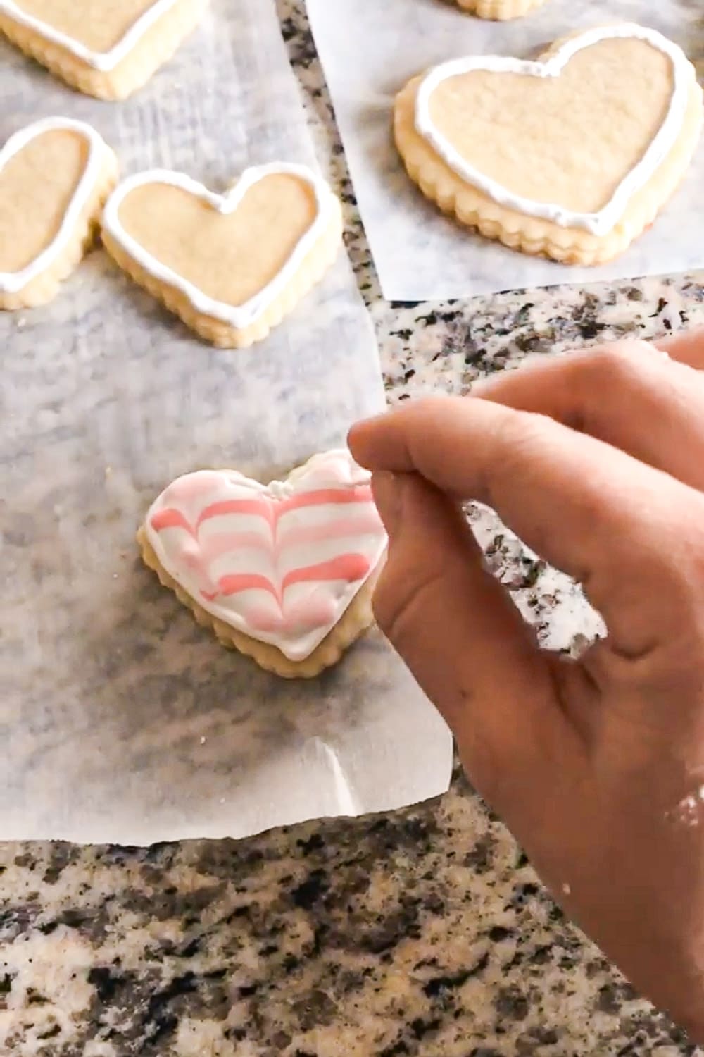 Using a toothpick to drag pink royal icing through white icing to create a feathered pattern on a heart-shaped cookie, with more outlined heart cookies behind it.