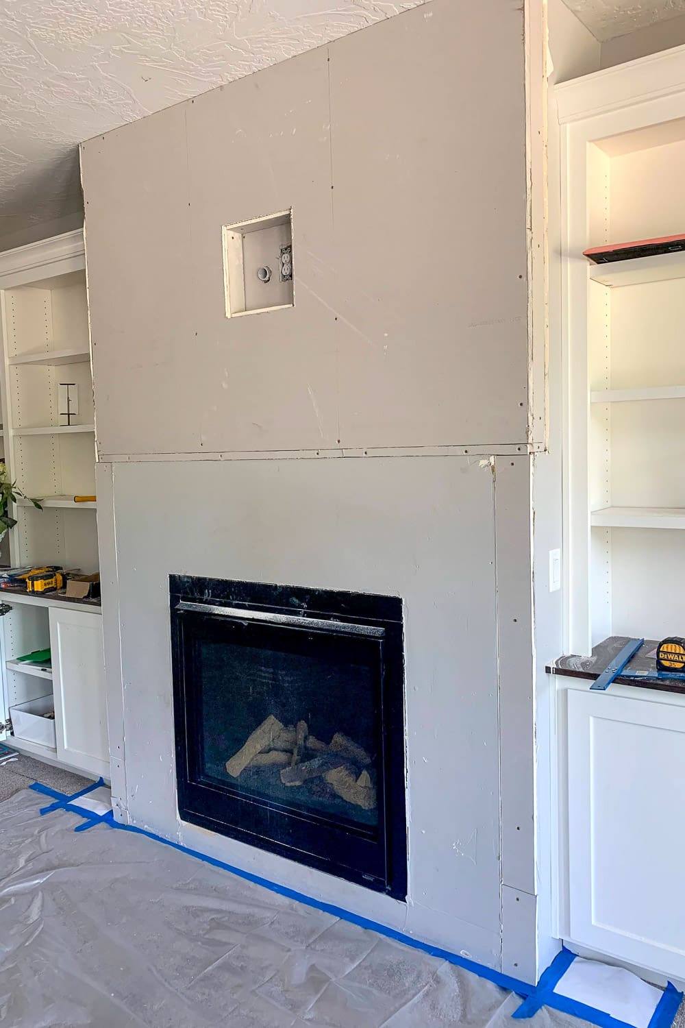 Fireplace wall with new sheetrock installed showing multiple pieces screwed in place, recessed media box cutout, plastic sheeting taped to floor