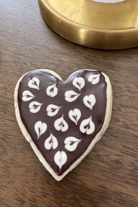 Heart-shaped sugar cookie flooded with dark royal icing without meringue powder and decorated with white piped heart designs on a wooden table.