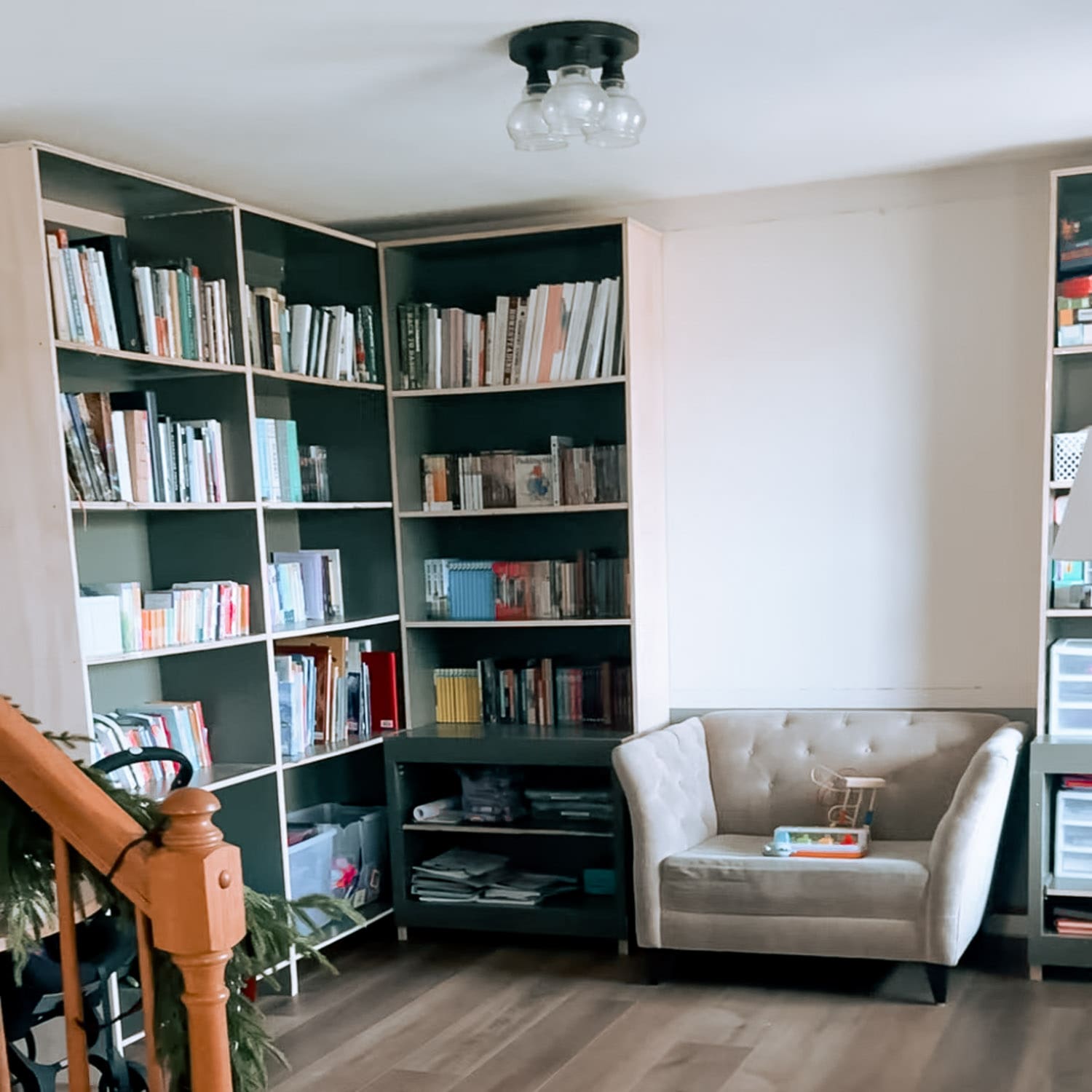 Wide view of our in-progress home library with DIY built-in bookshelves wrapping the corner, filled with books and homeschool supplies, with a cozy armchair and open floor space before painting.