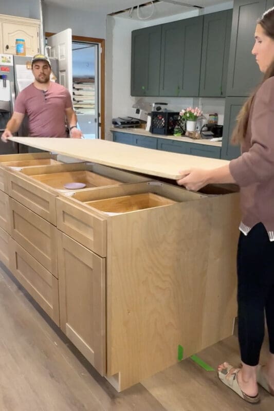 Setting the DIY kitchen island countertop in place by laying a plywood top onto the island cabinet base to test fit and prep for attaching the counter.