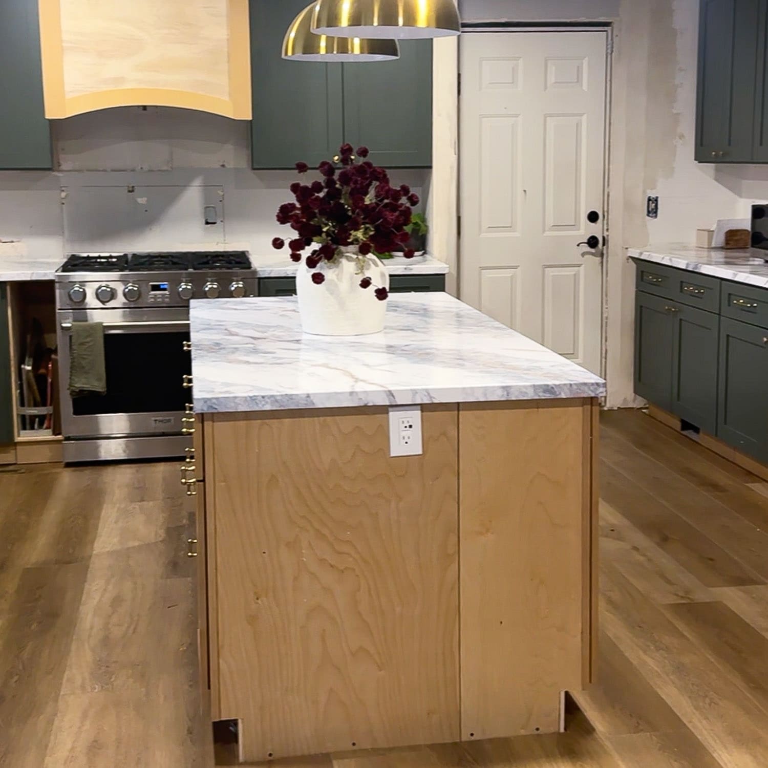 Wide view of our DIY kitchen island build in progress, showing an unfinished plywood island base topped with a white faux marble epoxy countertop, with green cabinets and brass pendant lights in the background.