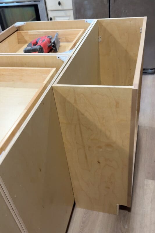 Close-up of the DIY kitchen island build showing the raw plywood cabinet base with an open end panel and drawer boxes installed, with a cordless nailer sitting on top during assembly.