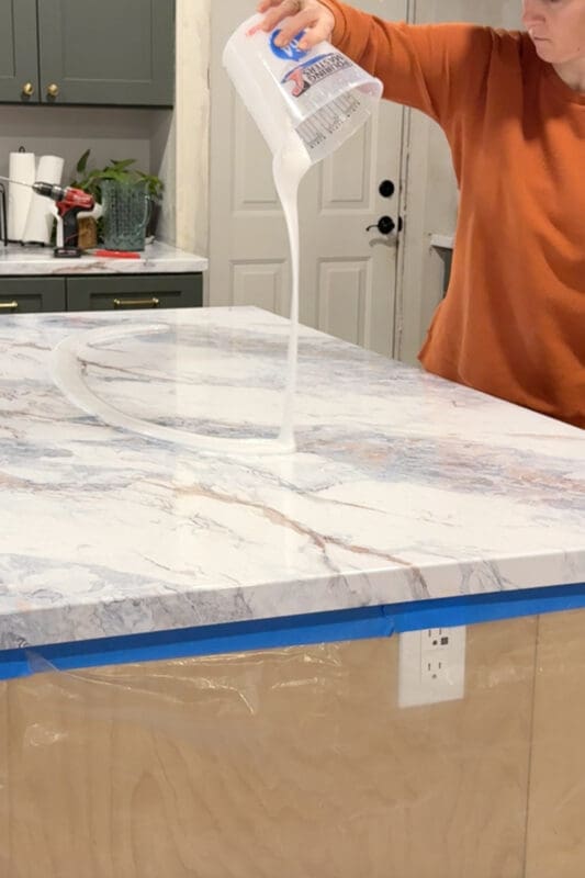 Pouring a clear epoxy coat over the marble-look contact paper on the countertop, with blue painter’s tape protecting the cabinet edges during the epoxy finish.