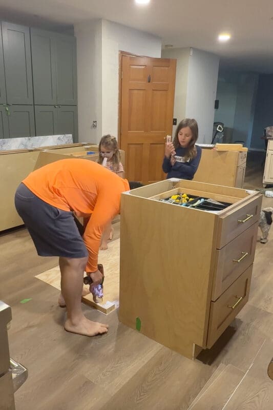 Installing DIY kitchen island base by screwing 2x4 blocking into the subfloor, creating a sturdy footprint for the island cabinets to sit on and be anchored in place.