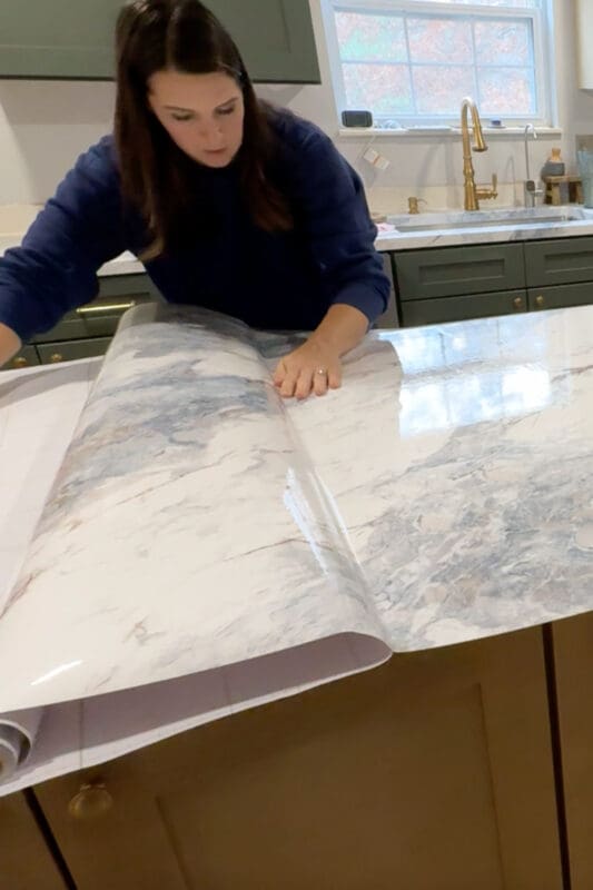 Applying marble-look contact paper to the countertop, smoothing the vinyl over the plywood top to create a faux stone surface before sealing.