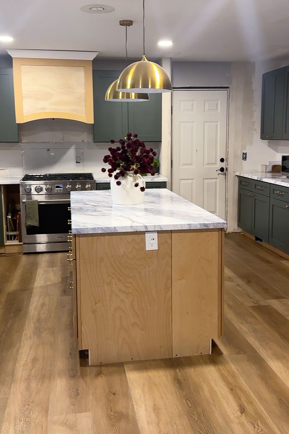 Closer look at the DIY kitchen island with a raw plywood cabinet base and a faux marble epoxy countertop, styled with a white vase of dark flowers under brass dome pendants in a green-cabinet kitchen.
