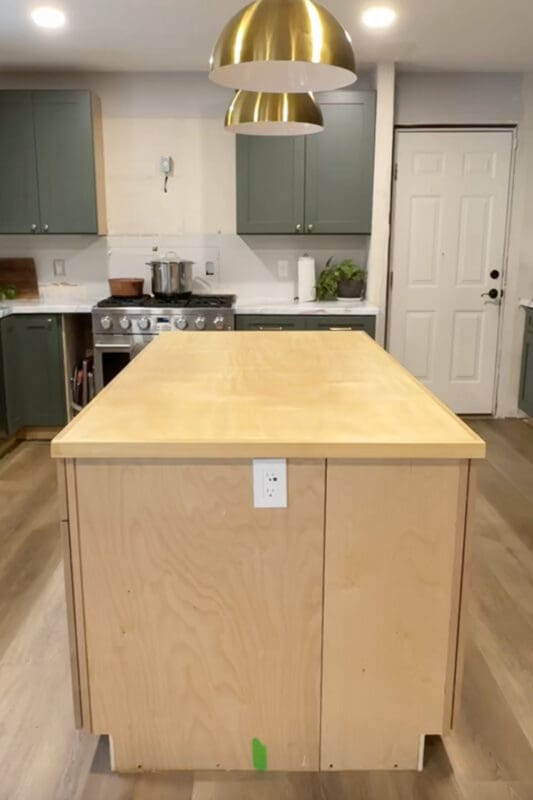 Front view of the DIY kitchen island with a raw plywood cabinet base and a plywood countertop installed, centered under brass pendant lights before finishing and painting.