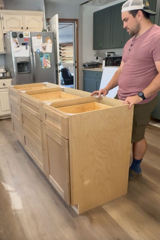 DIY kitchen island cabinet build in progress, showing a large unfinished plywood island with shaker-style drawer fronts being test fit in the kitchen before countertops and trim are installed.