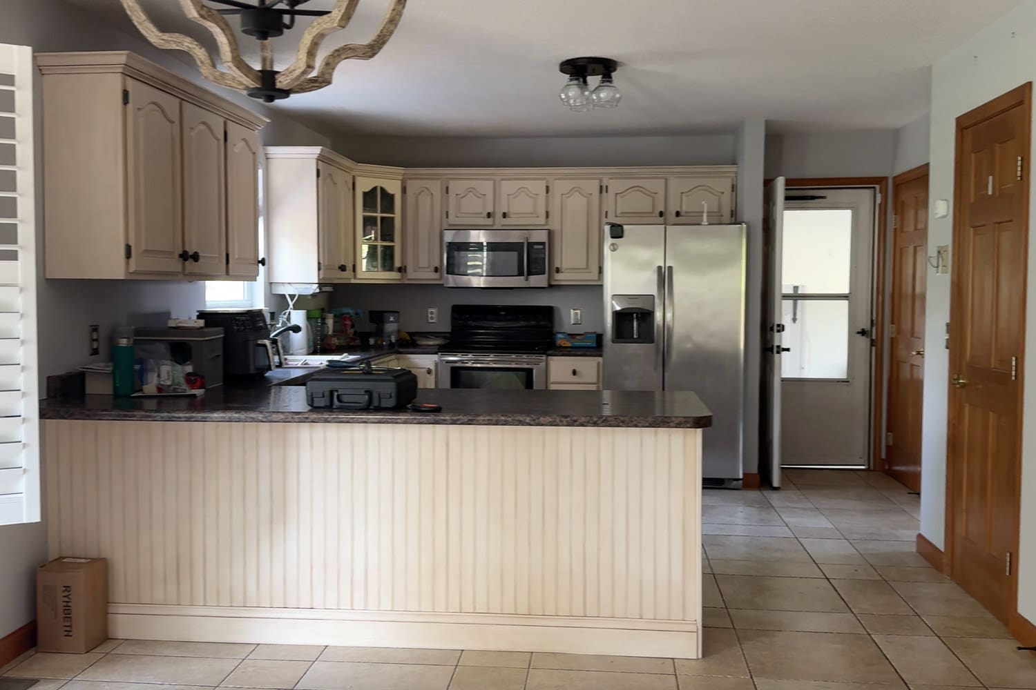 Before photo showing the original layout with builder-grade off-white cabinets, a raised bar peninsula, dark laminate countertops, tile flooring, and a stainless steel fridge.