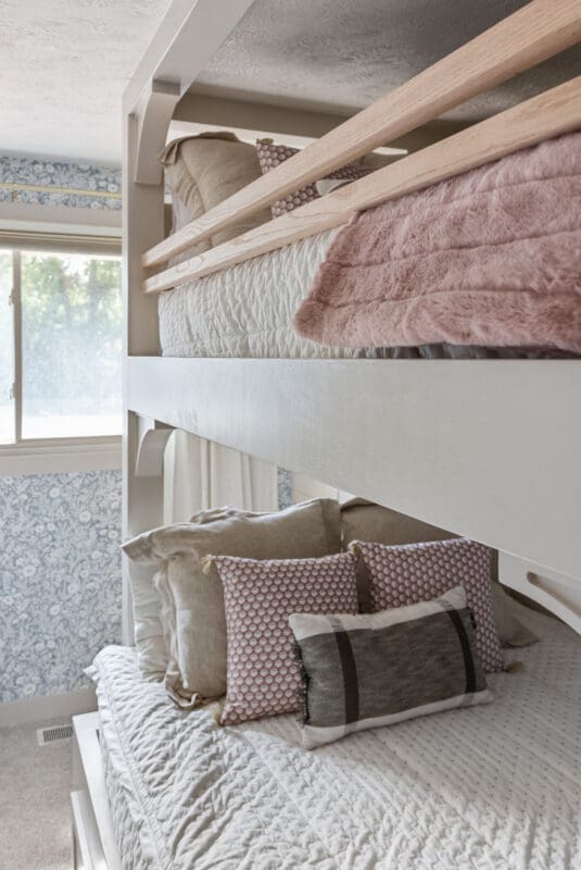Custom built-in full-over-queen bunk beds with white painted frame, natural wood guard rails, and cozy neutral bedding in a girls’ bedroom by a window.