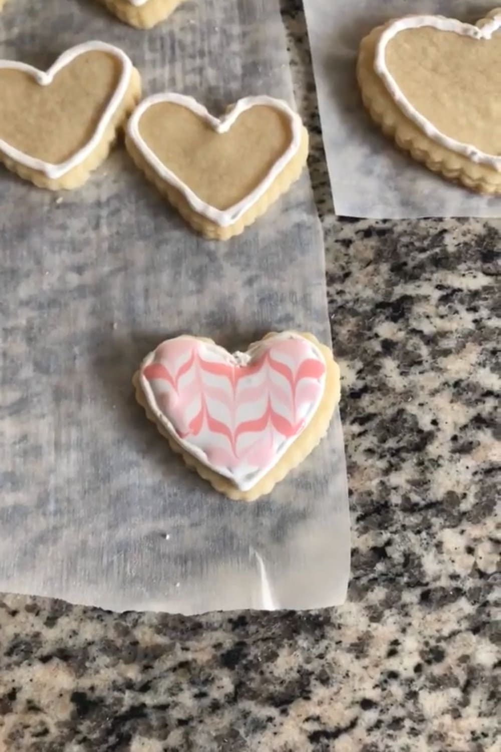 Heart-shaped sugar cookie decorated with a pink and white feathered royal icing pattern on parchment paper, with undecorated white-outlined heart cookies in the background.