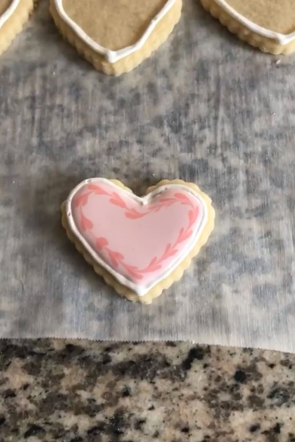 Finished heart-shaped sugar cookie flooded with light pink royal icing and a darker pink marbled border design, resting on parchment paper on a granite countertop.