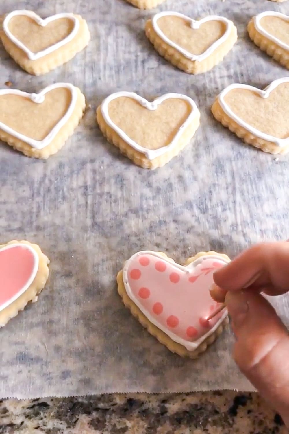 Decorating a heart-shaped sugar cookie with light pink royal icing, adding darker pink polka dots along the edge using a small piping tip on parchment paper.