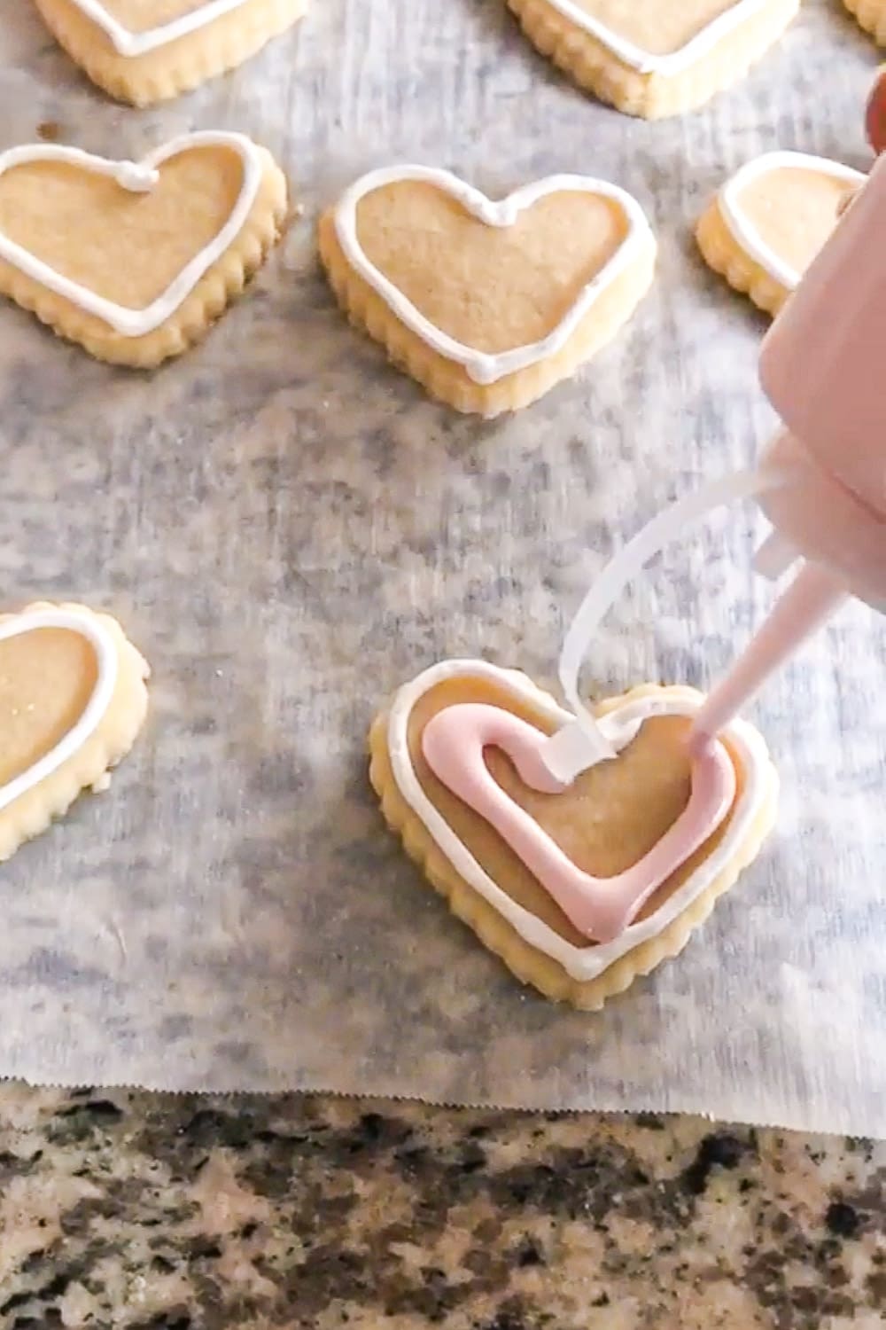 Close-up of heart-shaped sugar cookies on parchment paper as pink royal icing is piped inside a white outlined border for flooding.