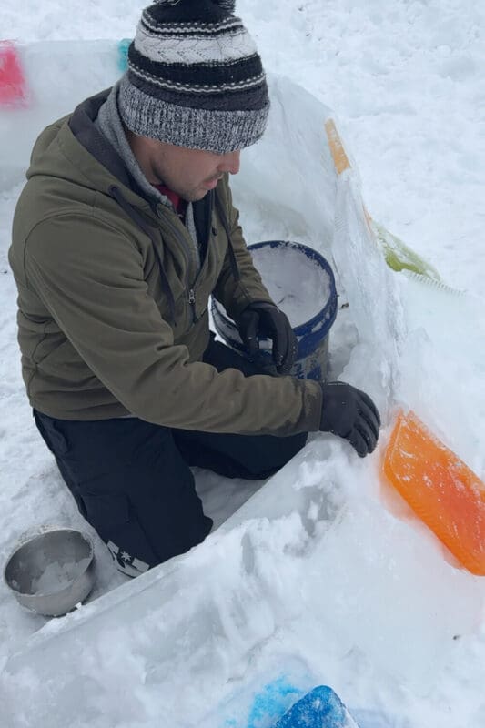 Applying snow-and-water slush mortar along the top of stacked ice blocks to seal the joints before adding the next layer in a how to build an igloo tutorial