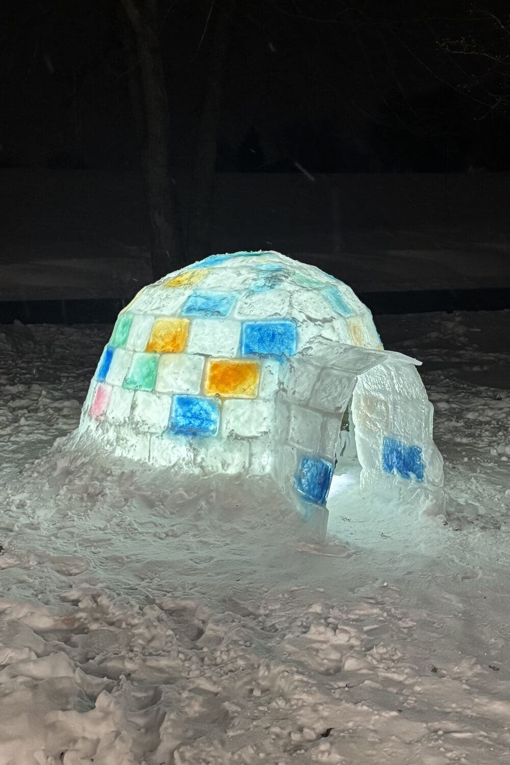 Finished DIY igloo at night with colored ice blocks and a glowing light inside, showing the arched entrance tunnel and snow-covered ground for a how to build an igloo tutorial