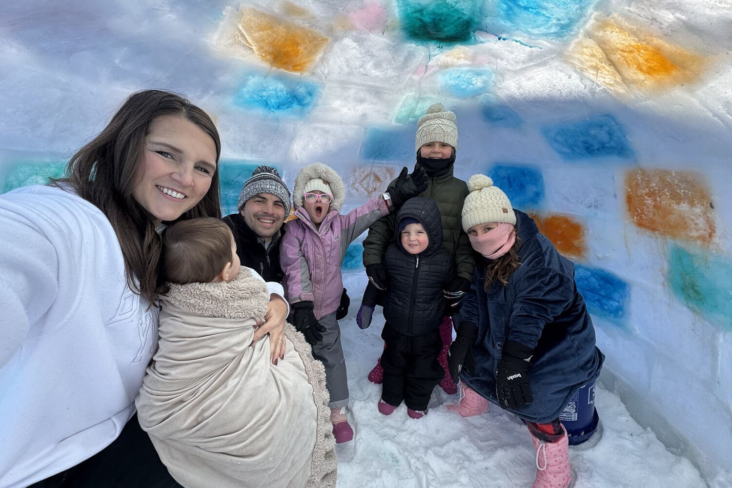 Family posing in front of a homemade backyard igloo built from ice blocks with colorful frozen panels, showing the finished winter project from a how to build an igloo guide